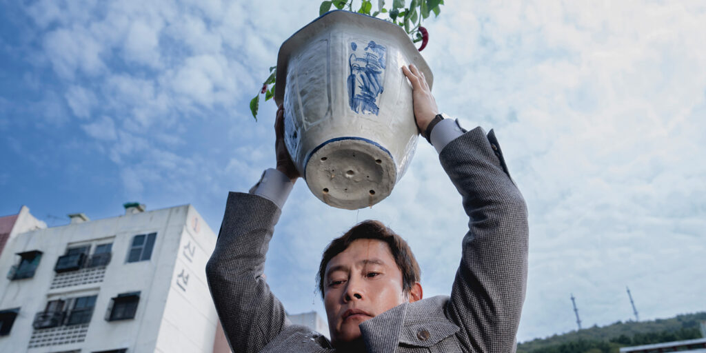 A man in a grey suit lifts a large ceramic flower pot above his head outdoors, with buildings, trees, and a blue sky with clouds in the background. Water drips from the bottom of the pot.