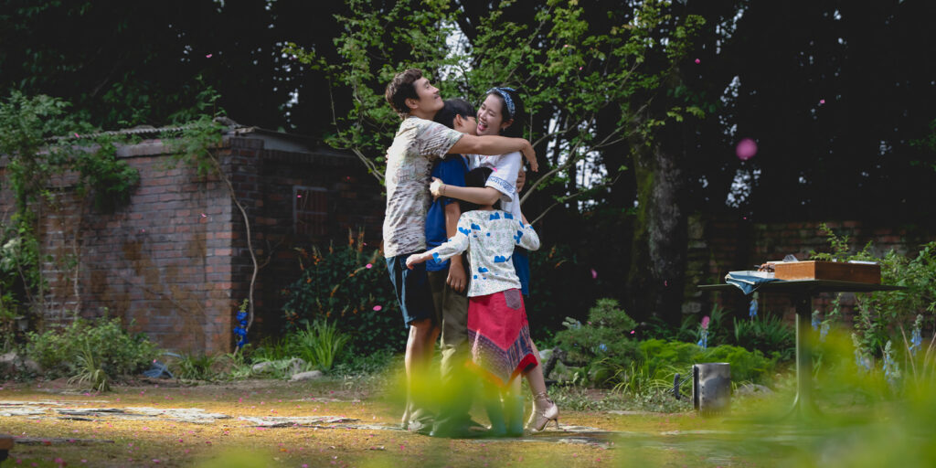 Three people stand outside in a garden hugging and smiling. Trees, plants, and a brick wall are in the background. A table with items is on the right side of the image.