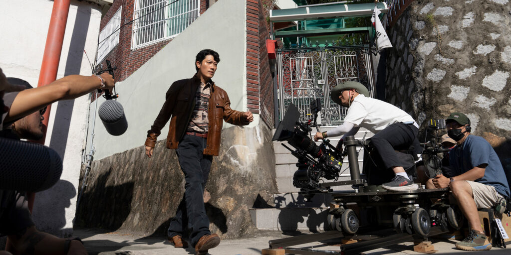 A man in a brown jacket walks past a film crew. One person holds a boom mic, and another films with a camera mounted on a track near stairs leading to a building.