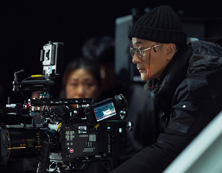 A person wearing a black beanie and glasses operates a large professional film camera on a set, whilst several other people stand in the background.