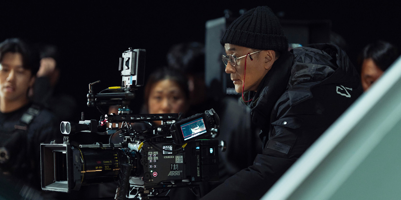 A person wearing a black beanie and glasses operates a large professional film camera on a set, whilst several other people stand in the background.