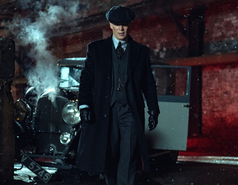 A man in a dark coat, suit, gloves, and flat cap walks away from a vintage car with smoke coming from its engine in an alley at night. Snow is falling, and red light illuminates the brick wall behind him.