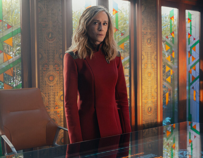 A woman in a long red coat stands behind a glass desk in a modern room with stained glass windows and patterned walls. She looks ahead with a neutral expression.