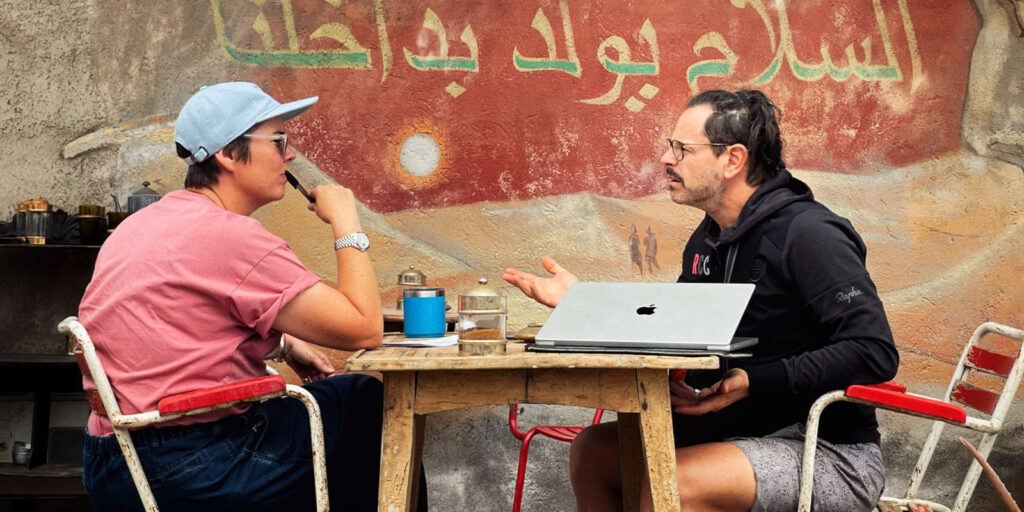 Two people sit opposite each other at a small wooden table outdoors, talking. One has a laptop open. Both have drinks. The background shows a worn wall with Arabic writing and faded artwork.
