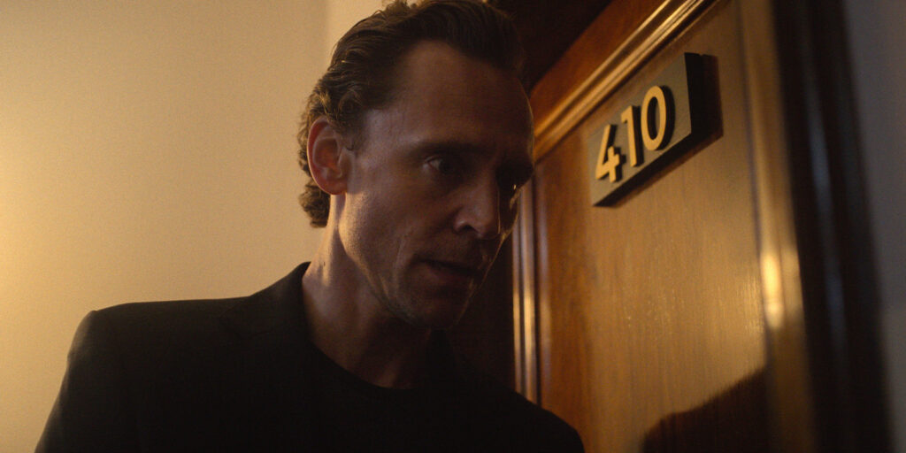 A man with short, dark hair stands in front of a closed wooden door marked with a gold “410” plaque. He is wearing a black shirt and jacket, looking intently to his right in dim lighting.