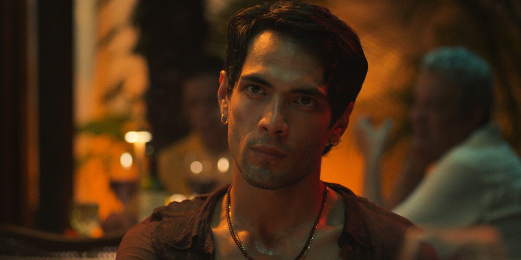 A man with short dark hair and facial stubble sits indoors, wearing a sleeveless top and a beaded necklace, looking forward with a serious expression. The background is dimly lit with blurred figures and warm lighting.