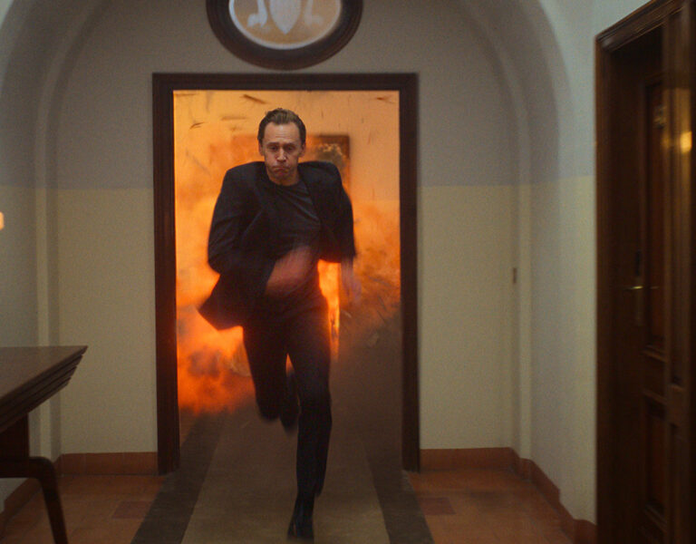A person in a suit runs down a corridor with a fiery explosion in the background, surrounded by closed doors and a table with lit lamps on the left side.