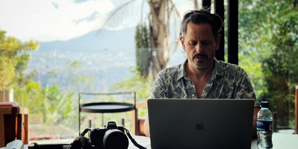 A man sits at a table working on a laptop with a camera, lens, and water bottle beside him. Large windows behind him show trees and an outdoor view with mountains in the distance.