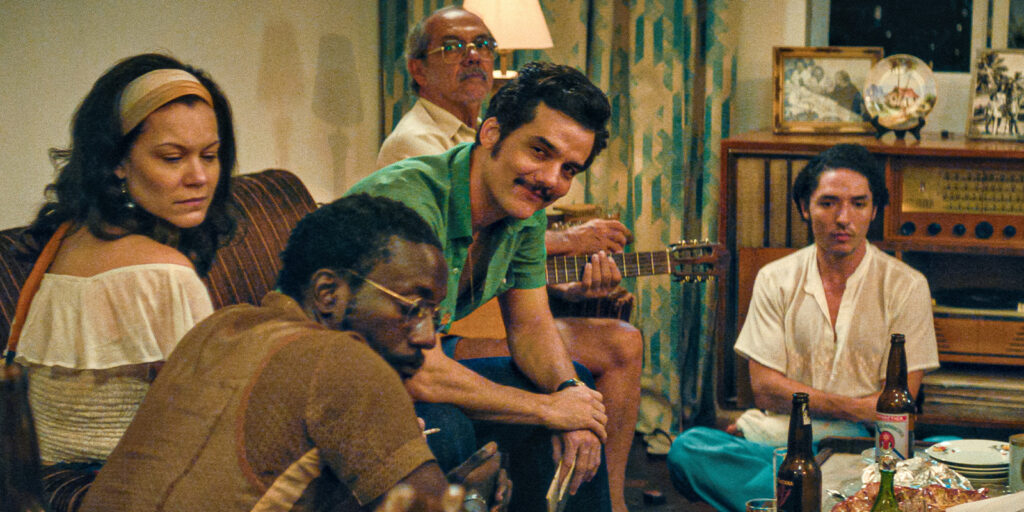 Five adults sit closely together in a warmly lit sitting room. One holds a guitar, and several have drinks. They appear relaxed amid homely decor, photographs, and a vintage radio in the background.