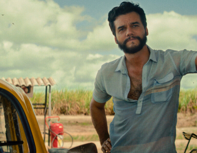 A man with dark hair and a beard stands beside a yellow car, resting one arm on an object out of frame. He is wearing a light blue short-sleeved shirt, and there is grass and a cloudy sky in the background.