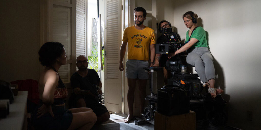 A film crew sets up a shot in a room with light coming through open doors; a woman sits on the floor whilst crew members operate a camera and equipment nearby.