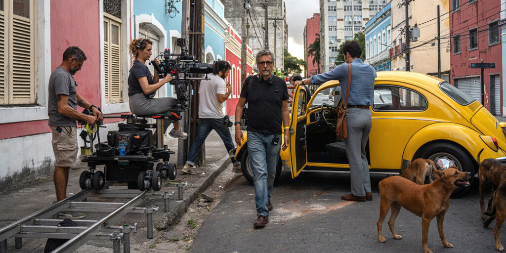A film crew shoots a scene on a city street. One person films with a camera on a dolly track, another checks equipment, and a man in smart clothes stands by a yellow Volkswagen Beetle. Several stray dogs are nearby.