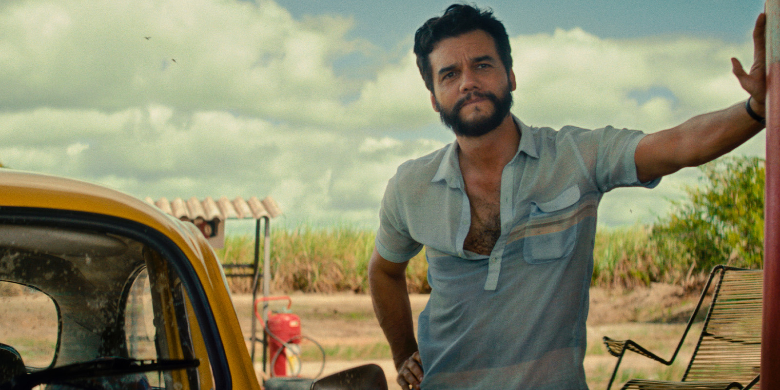 A man with dark hair and a beard stands beside a yellow car, resting one arm on an object out of frame. He is wearing a light blue short-sleeved shirt, and there is grass and a cloudy sky in the background.
