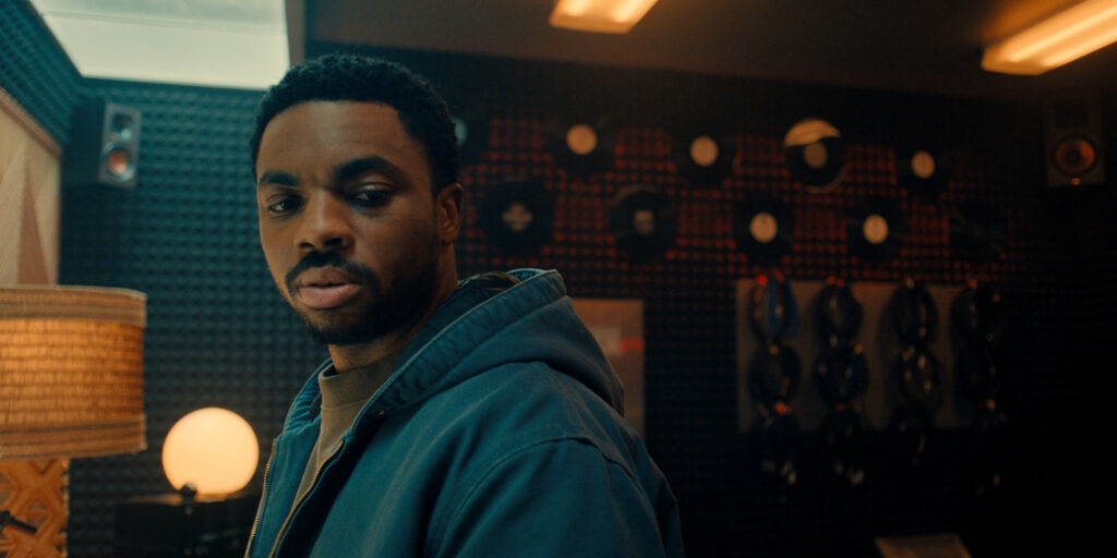 A man in a blue jacket stands in a dimly lit room with records on the wall and a lamp glowing on a table. He is looking off to the side with a neutral expression.