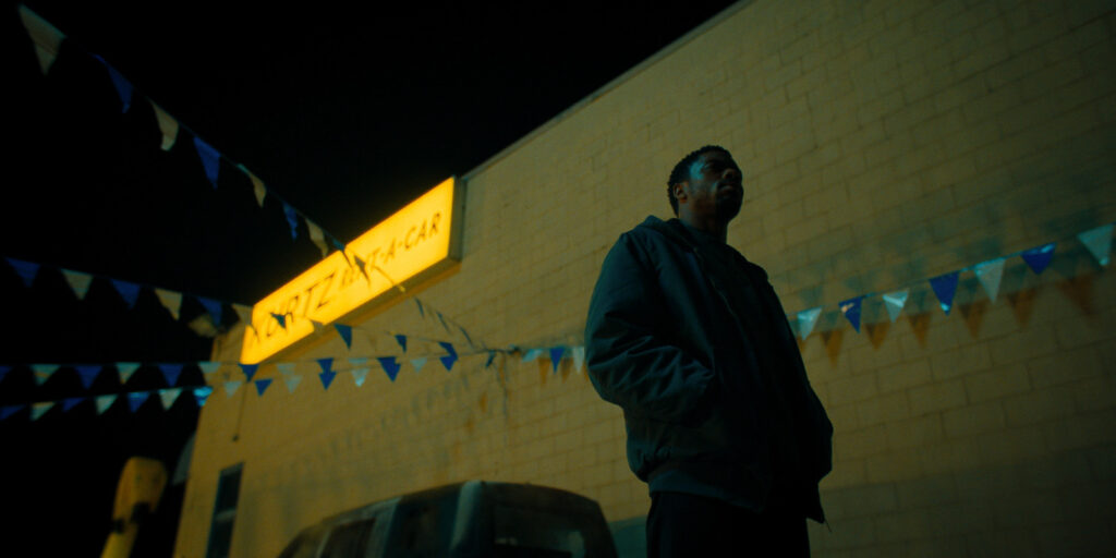 A person stands outside at night near a beige brick building with a glowing yellow sign that reads “MOTZA STAR CAR.” Blue and white pennant flags hang overhead. The person is in shadow, and a vehicle is partially visible.
