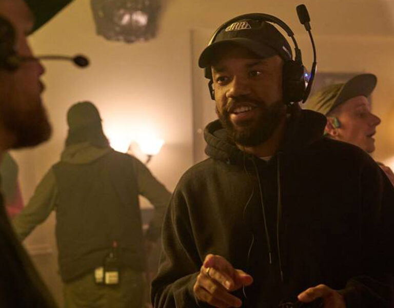 Several people wearing headsets and casual clothing work on a film or TV set, with lighting equipment and cameras visible in the background. One person in the foreground is speaking and gesturing.