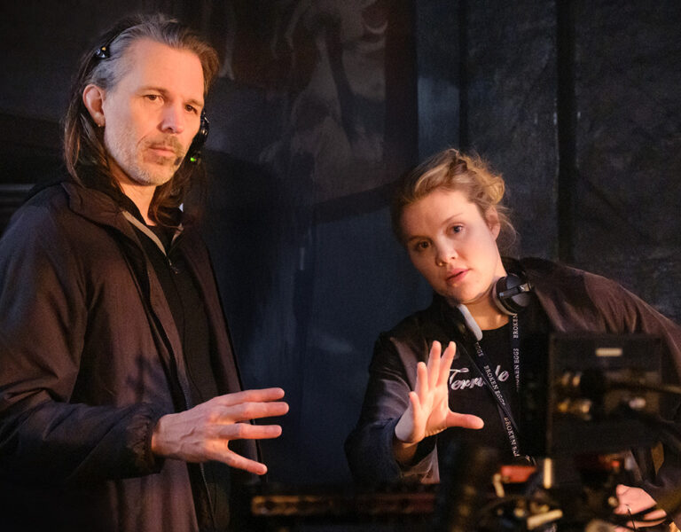 Two people wearing headsets and dark jackets stand indoors beside camera equipment, gesturing with their hands as they appear to discuss something during a film or TV production.