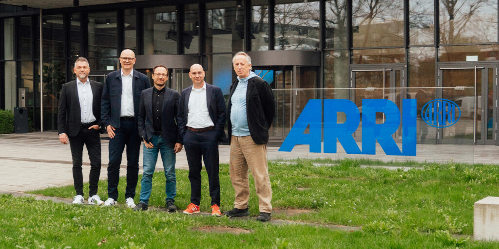 Five men stand outside the glass entrance of a modern building with a large blue ARRI logo on the window, next to a grassy area. They are dressed in smart casual attire.