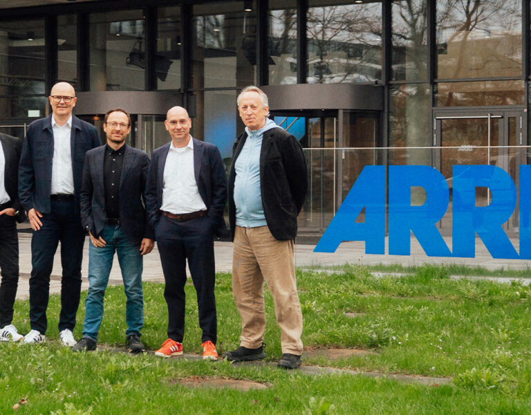 Five men stand outside the glass entrance of a modern building with a large blue ARRI logo on the window, next to a grassy area. They are dressed in smart casual attire.