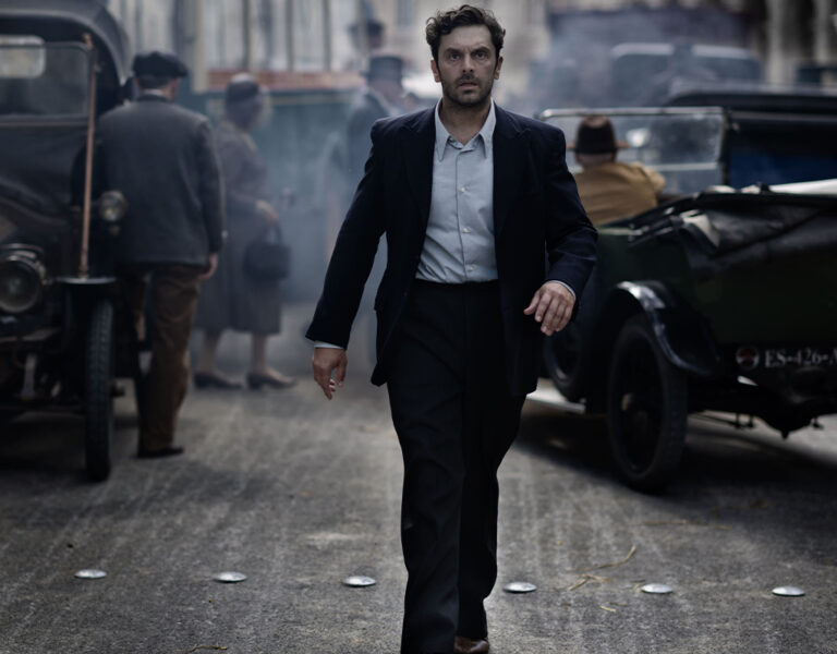 A man walking in a 1920s setting with cars in the background