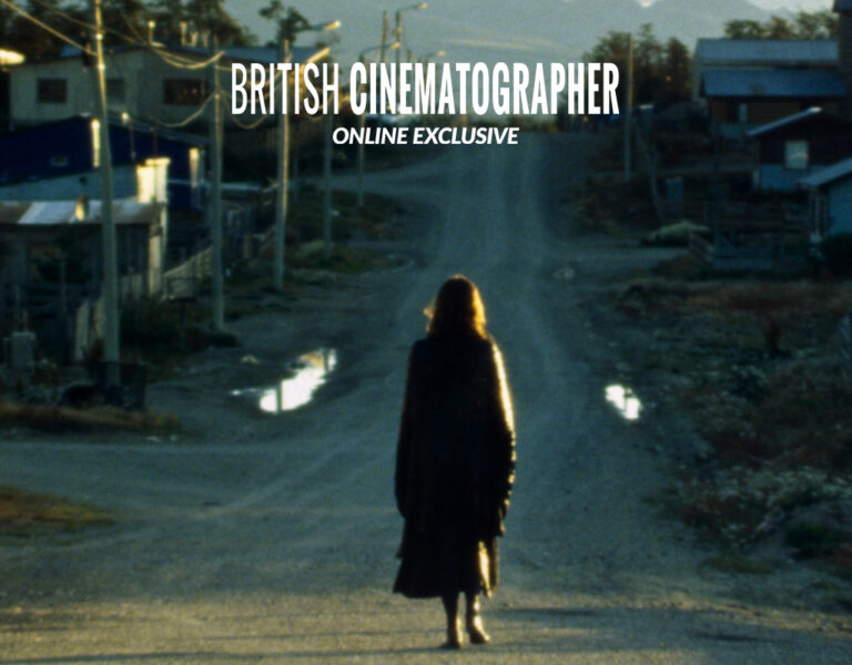 A person in a long coat walks alone down a dirt road towards a small rural town at dusk, with houses, telegraph poles, and mountains visible in the background. The words British Cinematographer Online Exclusive appear above.