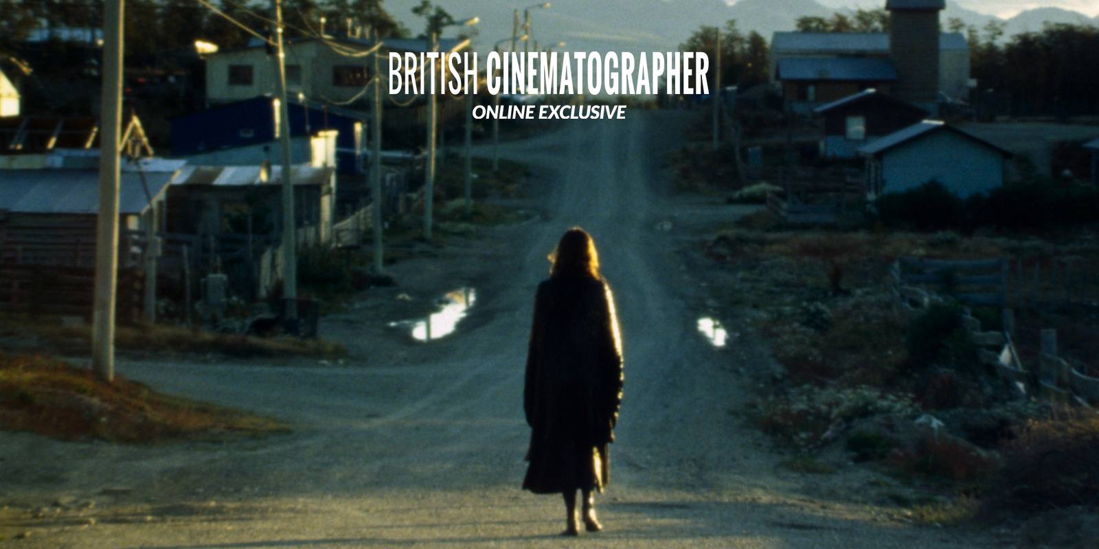 A person in a long coat walks alone down a dirt road towards a small rural town at dusk, with houses, telegraph poles, and mountains visible in the background. The words British Cinematographer Online Exclusive appear above.