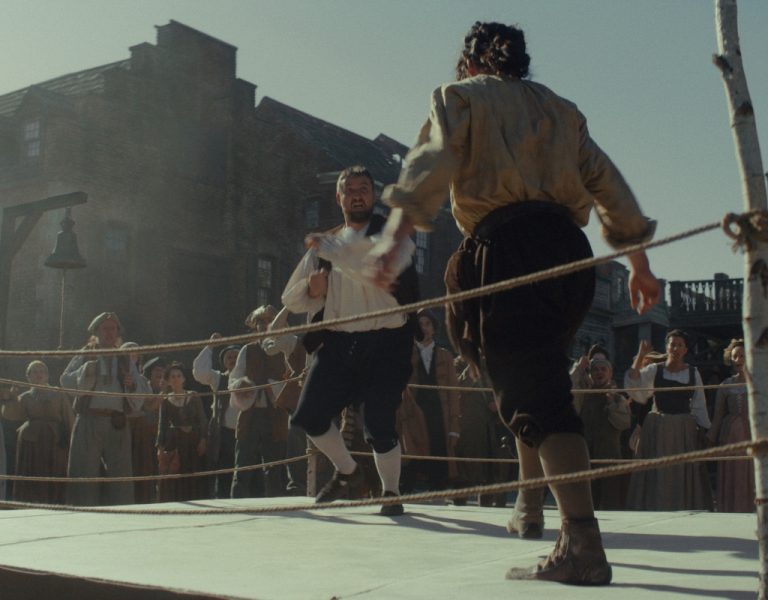 Two men in historical clothing are fighting in a roped ring outdoors. A crowd of people in period attire watches them. Old buildings are visible in the background. The scene appears to be set in the past.