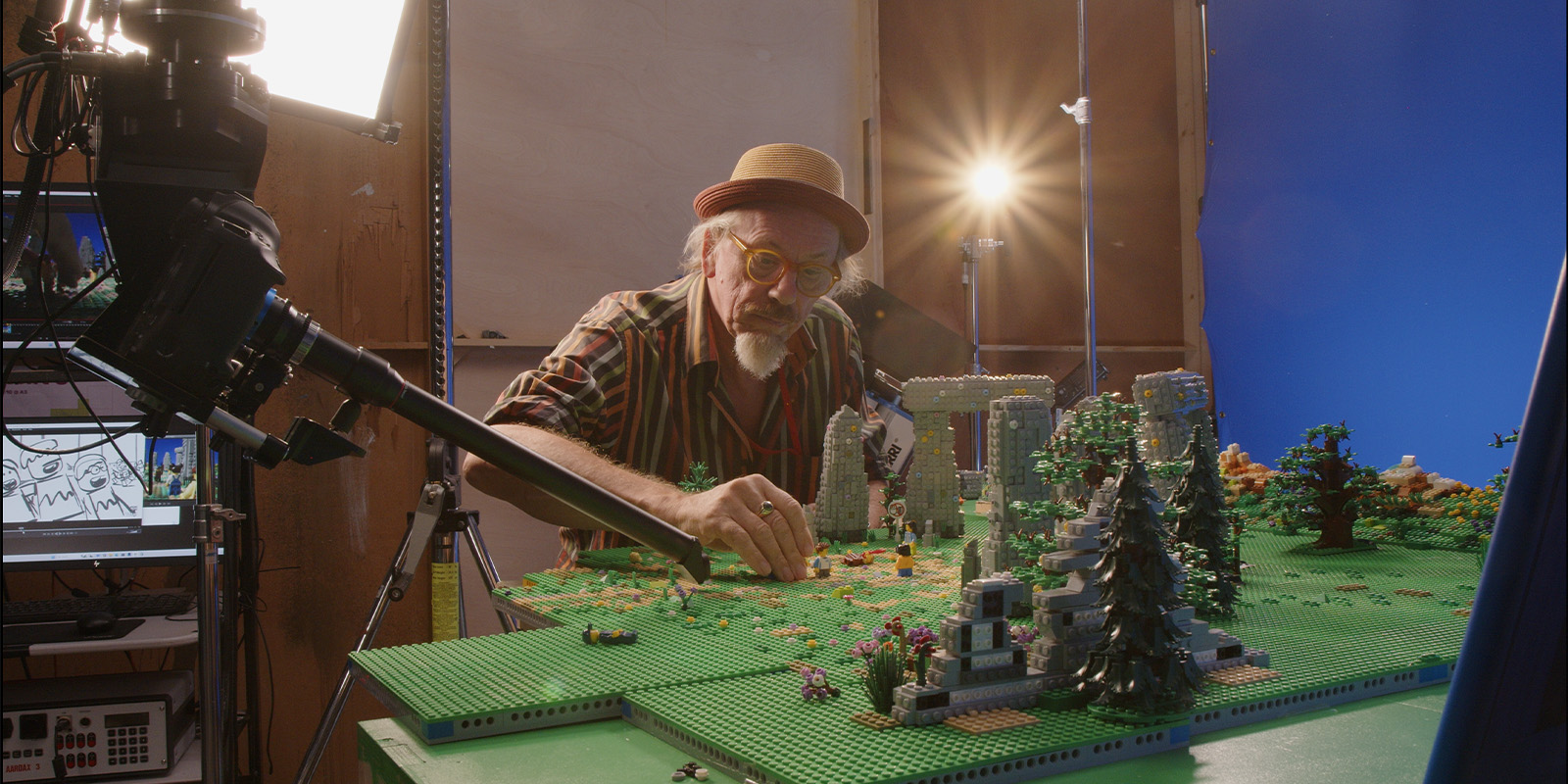 A person wearing a hat and glasses arranges pieces on a large, detailed LEGO set depicting a landscape, with professional cameras, lights, and a blue screen in the background.