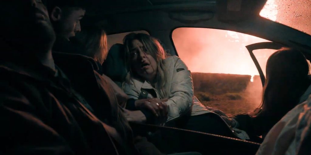 Four people sit inside a car at night; a woman in the front passenger seat looks distressed, holding hands with someone. A bright fire burns outside the car window, illuminating the interior with an orange glow.