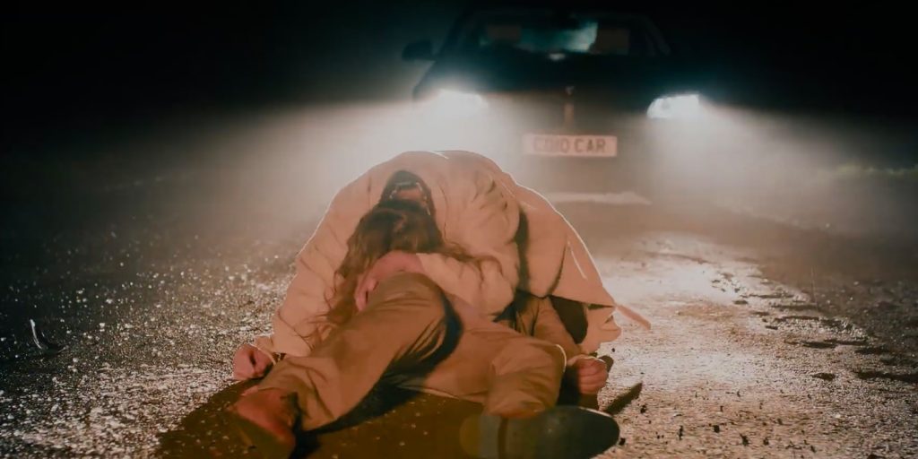Two people are on a wet road at night in front of a car with headlights on; one person, wearing a light coat, is hunched over another person lying motionless on the ground.