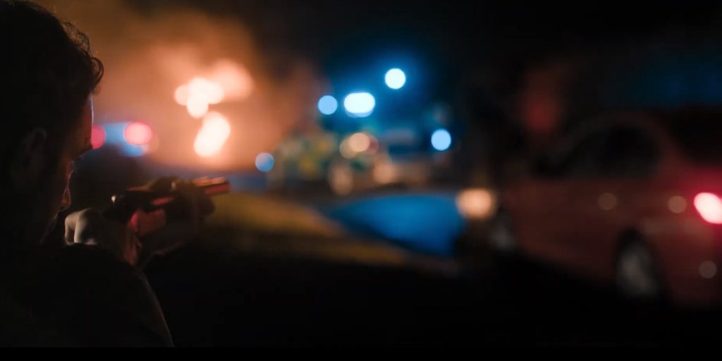 A person holding a shotgun aims towards a street scene at night, with cars and emergency vehicles in the background and a fire burning, casting an orange glow.