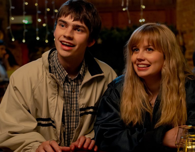 A young man and woman sit next to each other at a table in a warmly lit indoor setting. Both are smiling and looking slightly to their left. There are glasses and candles on the table in front of them.