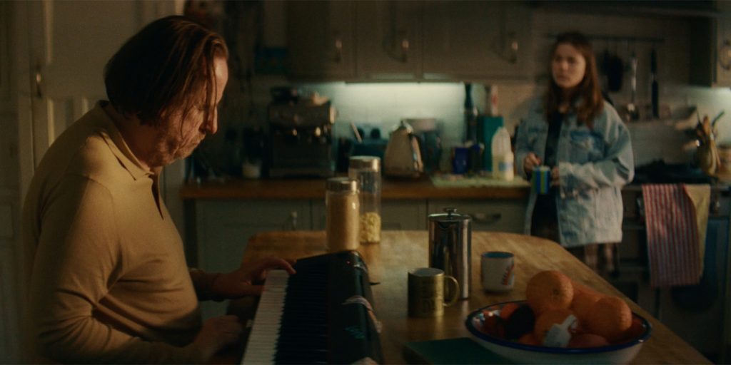A man plays an electric piano at a kitchen table, whilst a young woman stands in the background holding a cup. The kitchen worktop has a bowl of oranges, mugs, and various kitchen appliances.