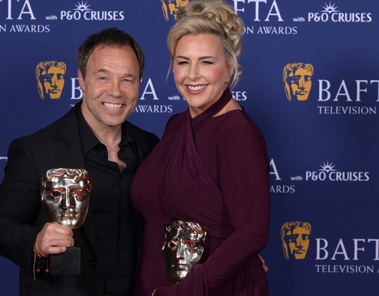 Two people posing on a red carpet holding awards