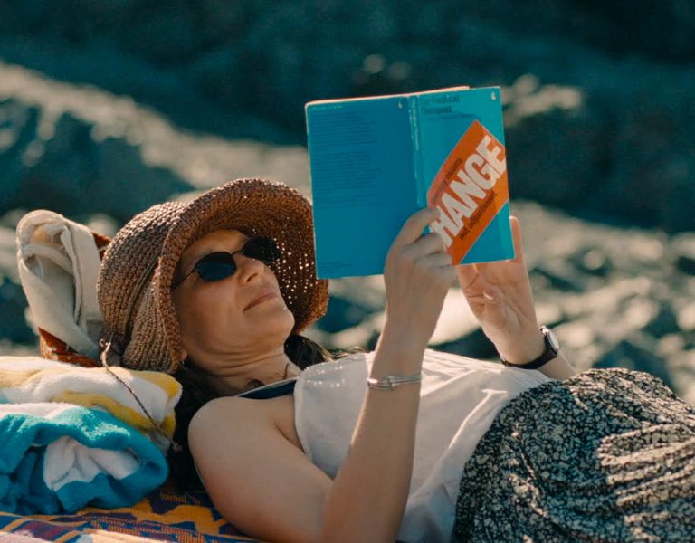 A woman wearing a sunhat, sunglasses, and a vest lies on a blanket outdoors, reading a blue book titled CHANGE with a towel and rocky background nearby.
