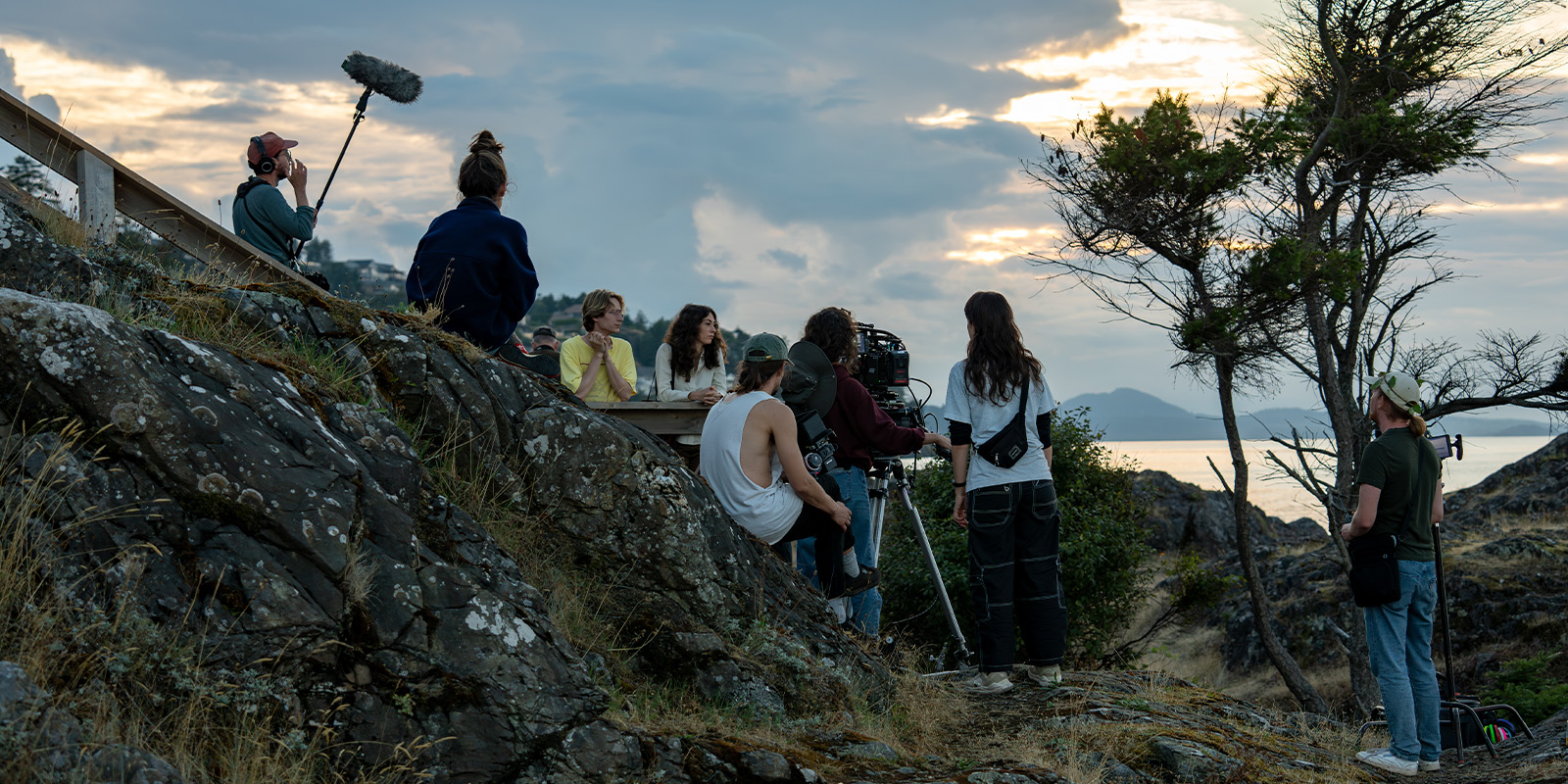A film crew is gathered on a rocky hillside at sunset, with some people seated near a railing and others around a camera on a tripod. One person holds a boom microphone, and another stands beside a tree overlooking the water.
