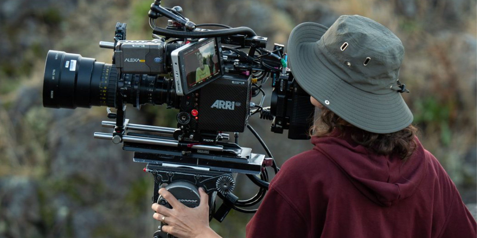 A person wearing a wide-brimmed hat and maroon hoodie operates a large ARRI Alexa cinema camera outdoors, focusing on the camera’s monitor with rocky terrain in the background.