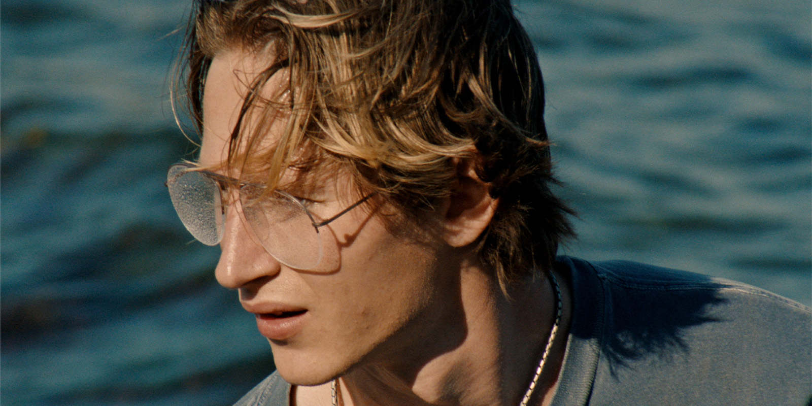 A young person with tousled blonde hair and clear-framed glasses looks to the side, standing near a body of water. They are wearing a blue shirt and a necklace.