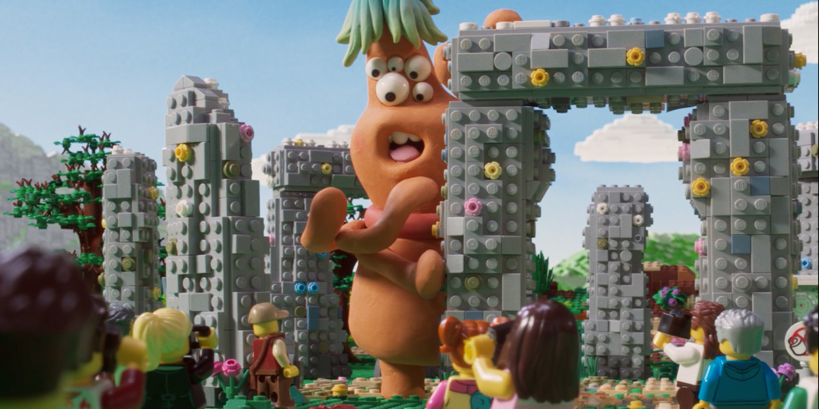 A large, orange, cartoonish creature with green hair stands among stone-like archways made of LEGO bricks, whilst a group of LEGO minifigures watch in the foreground under a blue sky.