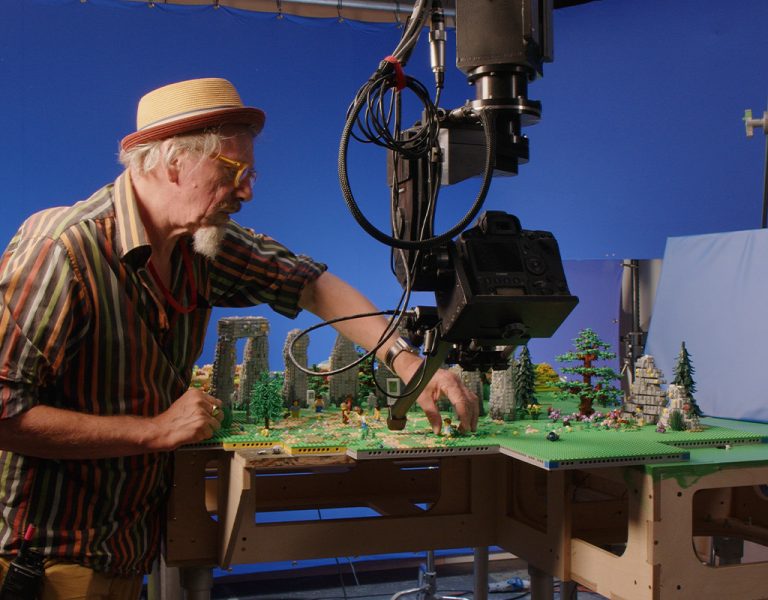 An older man in a striped shirt and hat arranges figures on a detailed miniature set in front of a camera rig, with a blue screen background and studio lighting.