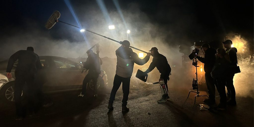 A film crew works at night on a smoky outdoor set, with a car, lighting equipment, and a boom microphone visible. Several crew members are adjusting equipment and filming in low light conditions.