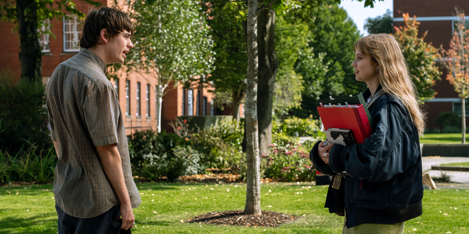 A young man and a young woman stand facing each other on a grassy campus with trees and buildings in the background. The woman holds several folders and notebooks. Both appear to be having a conversation outdoors.