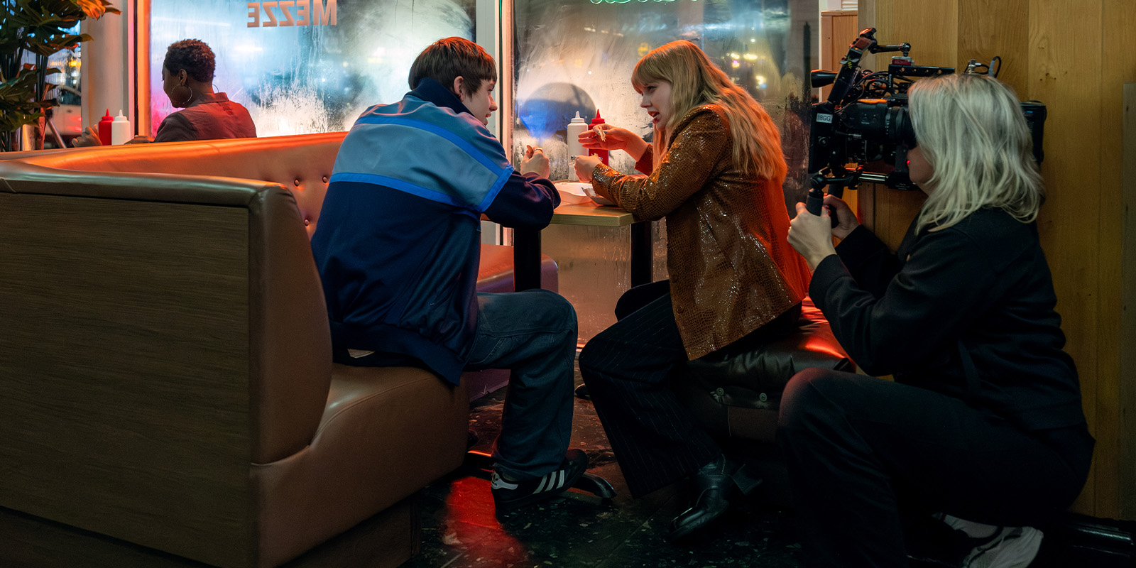 A camera operator films a young man and woman sitting opposite each other in a diner booth at night. They are engaged in conversation with drinks on the table. Another person sits alone at a nearby booth.