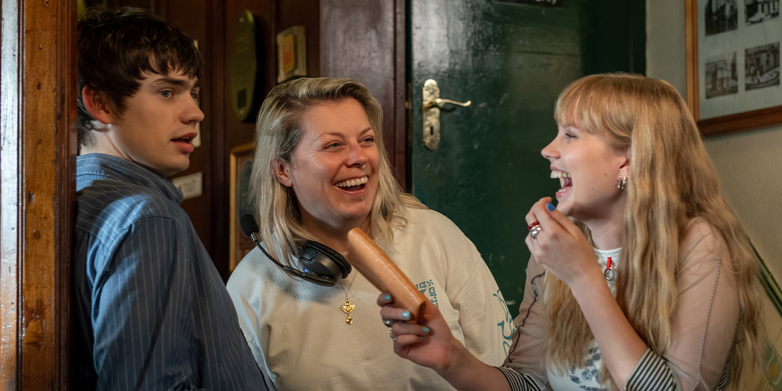 Three people stand indoors; a young man leans against a wall, a woman in the middle laughs, and a young woman on the right smiles whilst holding a peeled banana towards the young man.