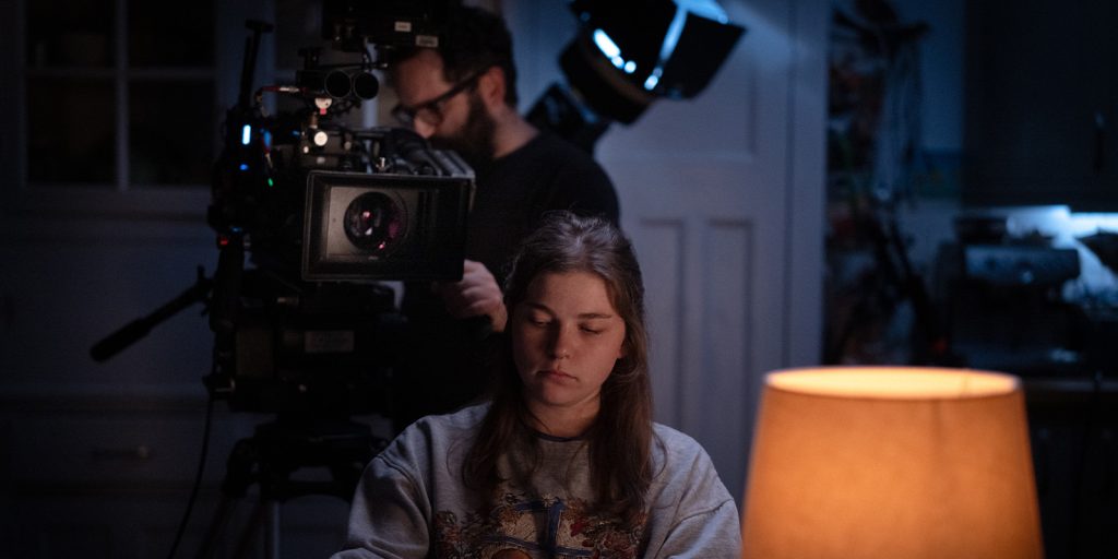 A young woman sits in front of a lit table lamp, looking down with a neutral expression. Behind her, a person operates a large film camera, and professional lighting equipment is visible in the dim room.