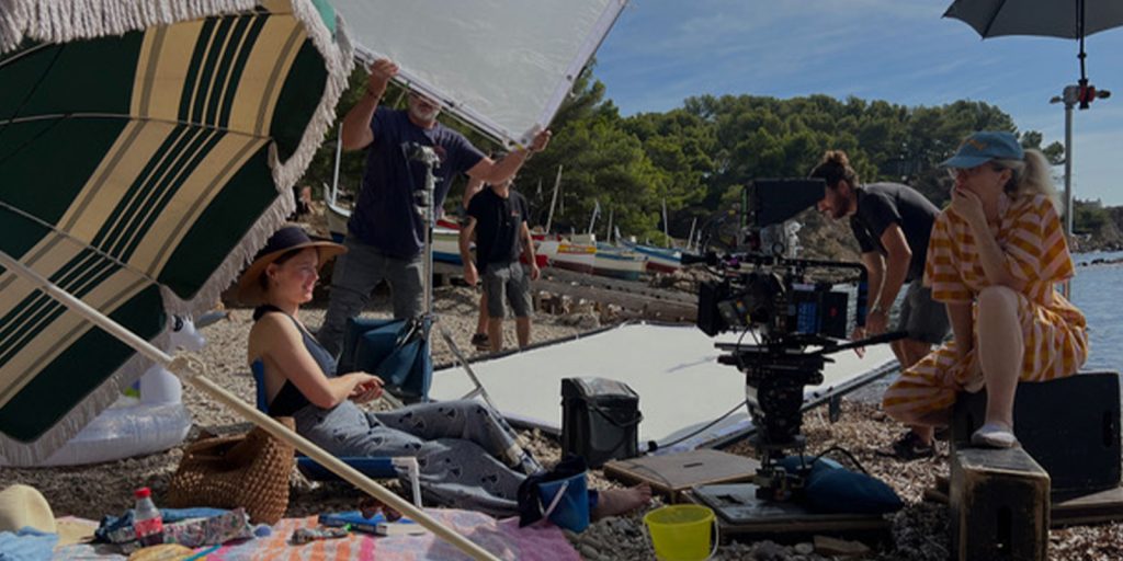 Two people sit under a parasol on a stony beach surrounded by film equipment, while three crew members hold reflectors and a camera is set up on a tripod. Boats and trees are visible in the background.