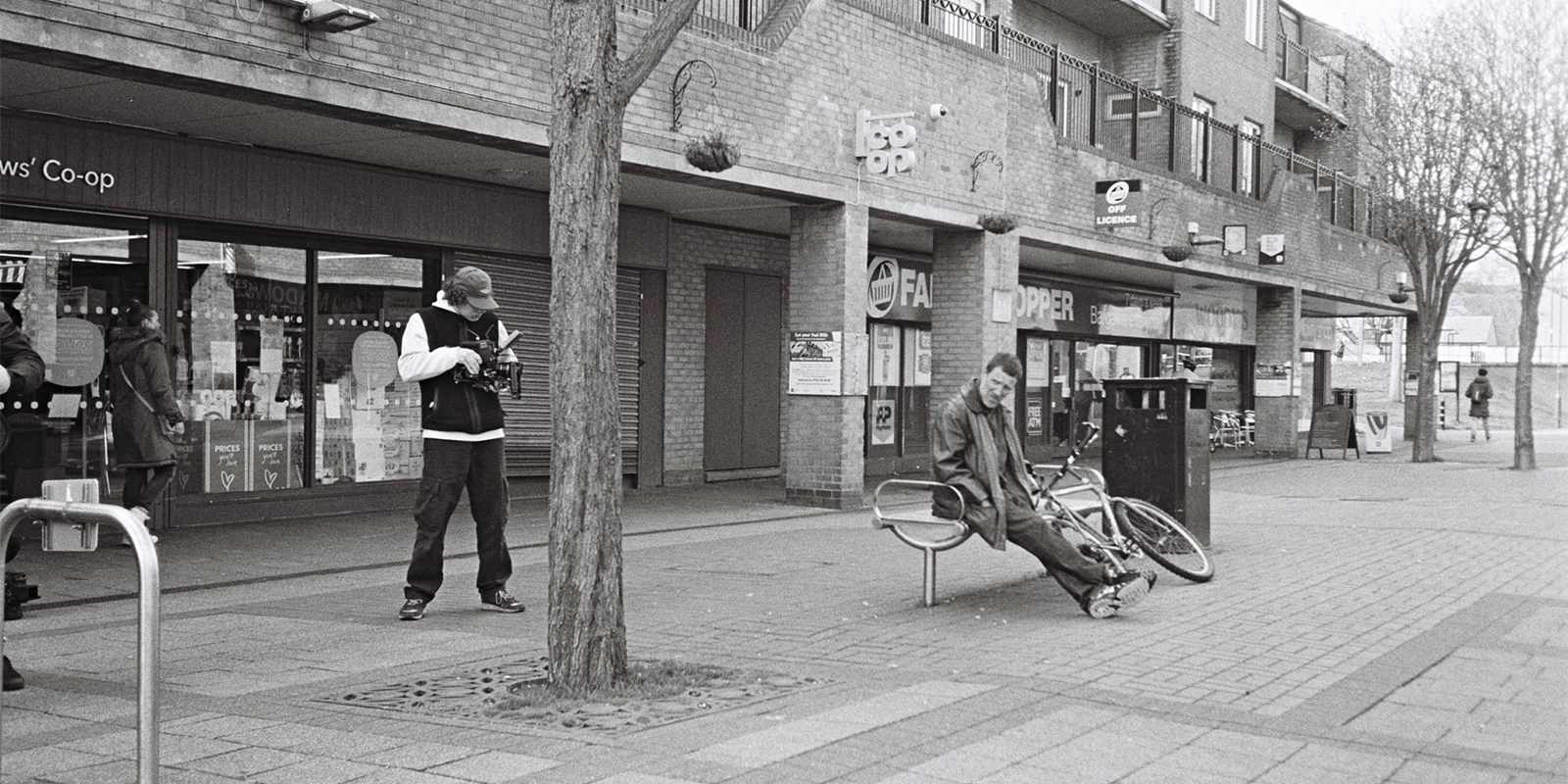 A man filming another man who is sat on a bench