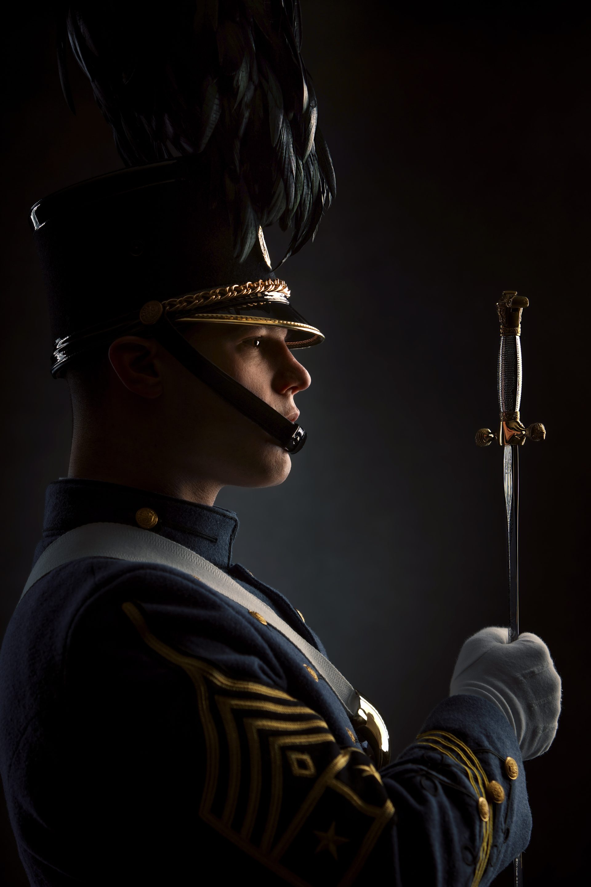 A person in a formal military-style uniform with a feathered hat holds a sword upright. The uniform features white gloves, decorative buttons, and a shoulder strap. The image is lit dramatically from the side.