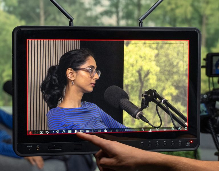 A person in a blue striped shirt sits in front of a microphone whilst being recorded; a camera operator views the scene on a large monitor, with trees visible through a window in the background.