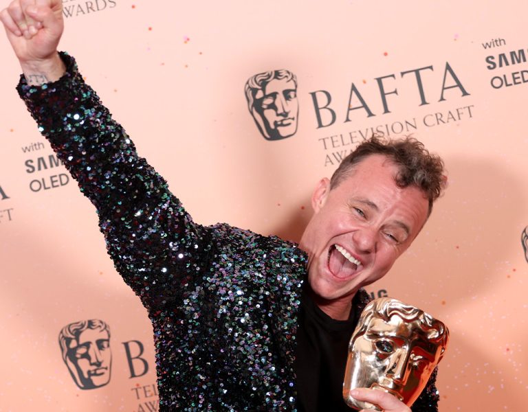 A person wearing a sequinned jacket raises one arm and smiles whilst holding a BAFTA trophy against a backdrop with BAFTA Television Craft Awards and Samsung OLED logos.